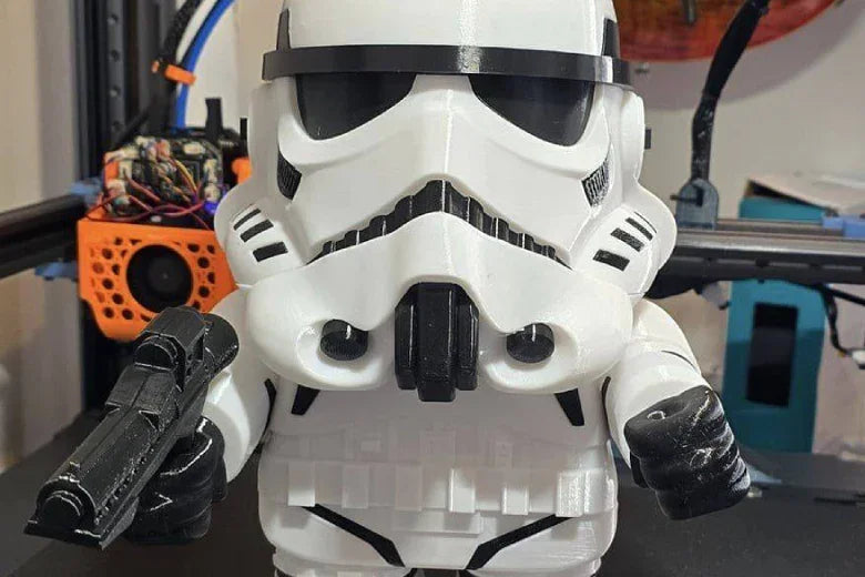 3D printed chibi stormtrooper figurine holding blaster, displayed in front of 3D printer